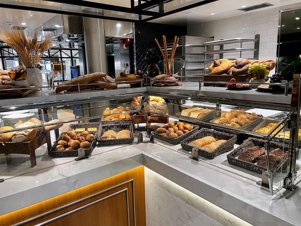 A bakery display case on the Costa Toscana, featuring a variety of breads, rolls, and pastries arranged in woven baskets on a multi-tiered marble counter.
