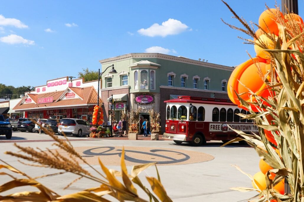 A red and white trolley parked in front of downtown storefronts during a Branson getaway, with pedestrians on the sidewalk and orange decorative balloons and corn stalks in the foreground.