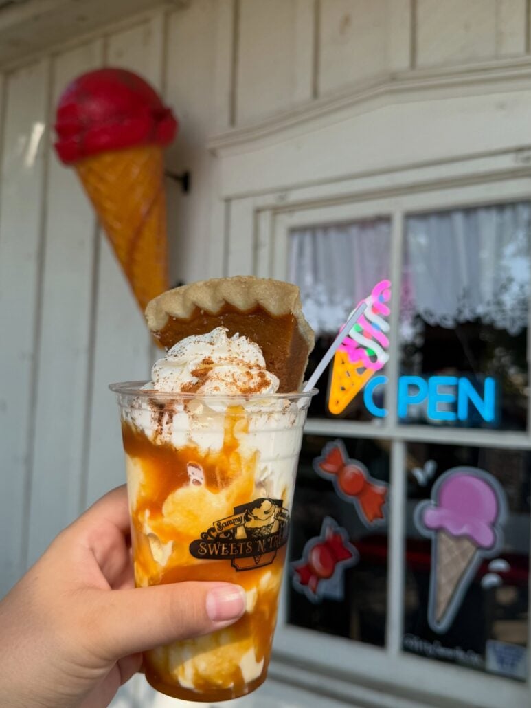 A hand holds a caramel-drizzled ice cream sundae topped with whipped cream and a slice of pie in front of an ice cream shop, the perfect treat to include when you plan a Branson trip.