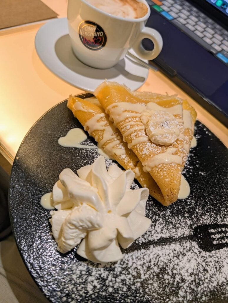 Two folded crepes drizzled with sauce and topped with a banana slice, served with a swirl of whipped cream on a black plate next to a cup of coffee.