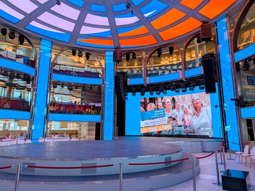 An indoor circular performance stage surrounded by a large digital screen displaying an advertisement for a land excursion package.