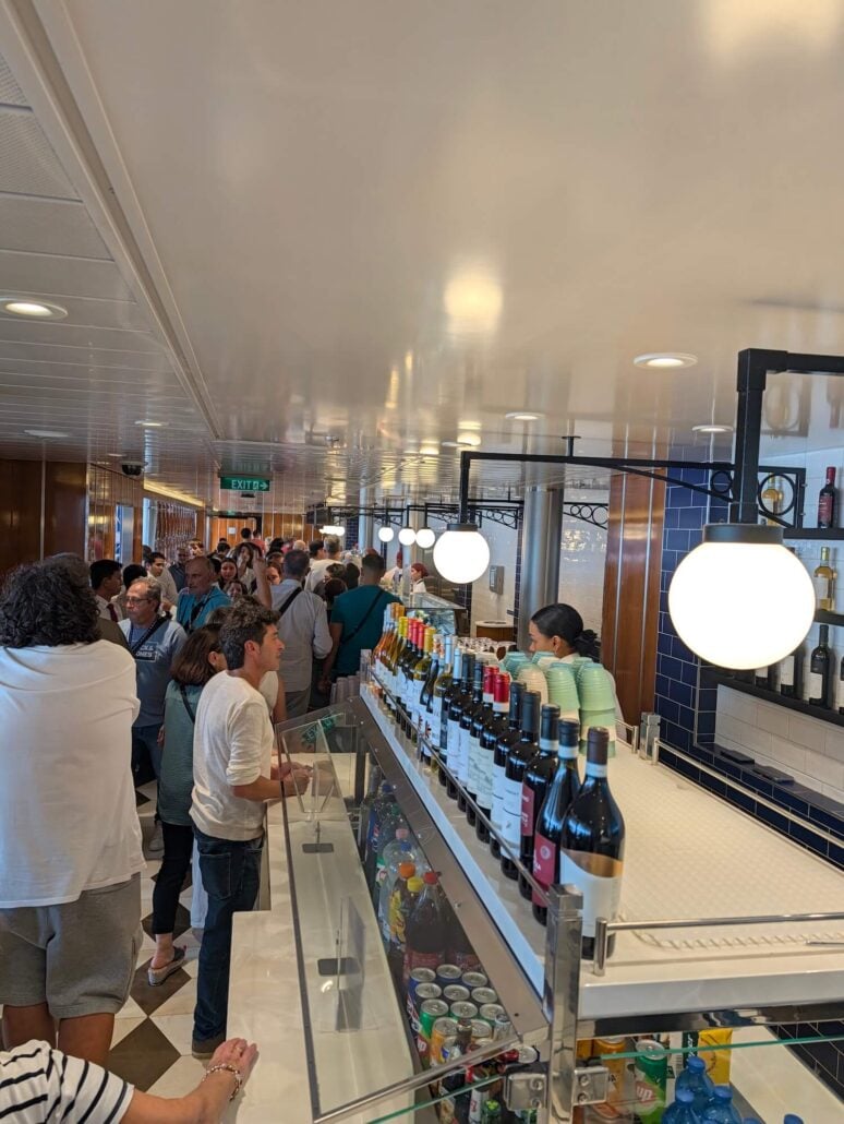 A crowded cruise ship bar area with customers lining up in front of a counter displaying bottles of wine and refrigerated drinks.