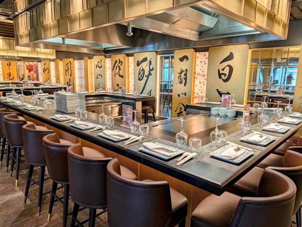A teppanyaki restaurant counter with brown stools and place settings arranged around a flat-top grill, featuring decorative panels with large black calligraphy on the wall behind.
