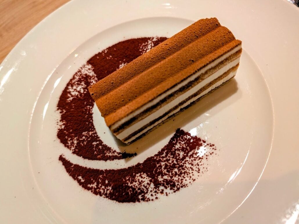A rectangular layered cake dusted with cocoa powder on a white plate with a decorative swirl of cocoa.
