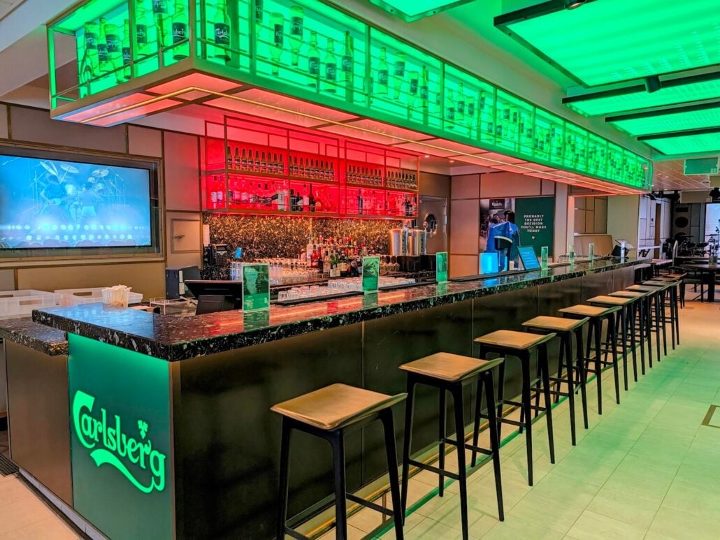 A modern bar with a black marble counter, high stools, and shelves of bottles illuminated by vibrant green and red neon lighting, featuring the Carlsberg logo on the front panel.