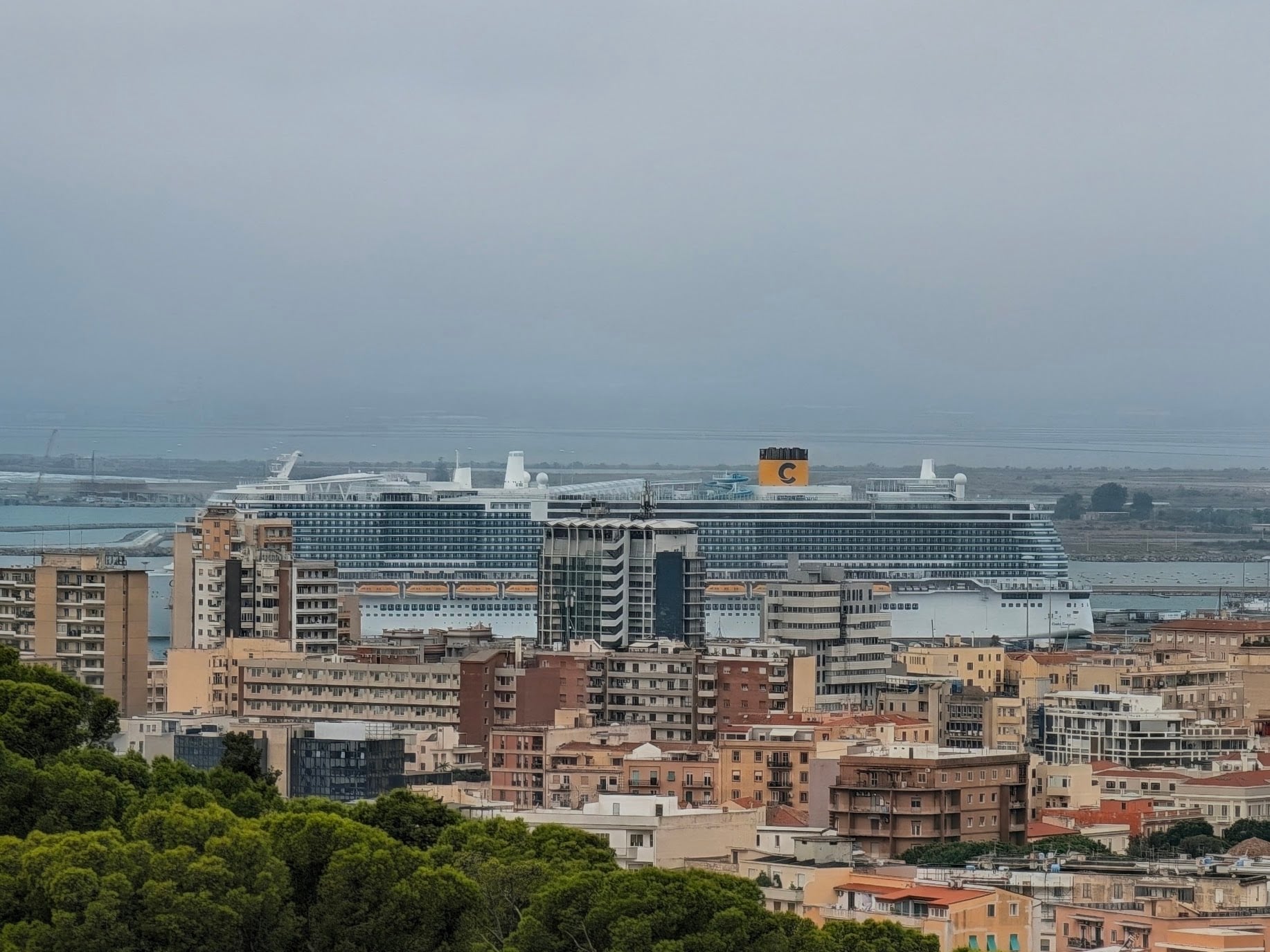 A Costa Toscana cruise ship with a yellow funnel docked behind a dense urban cityscape.