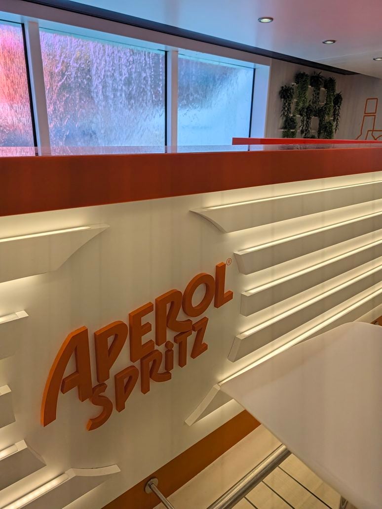 A white bar counter with a large orange “Aperol Spritz” logo, situated in front of a window with a water feature on the Costa Toscana cruise ship.