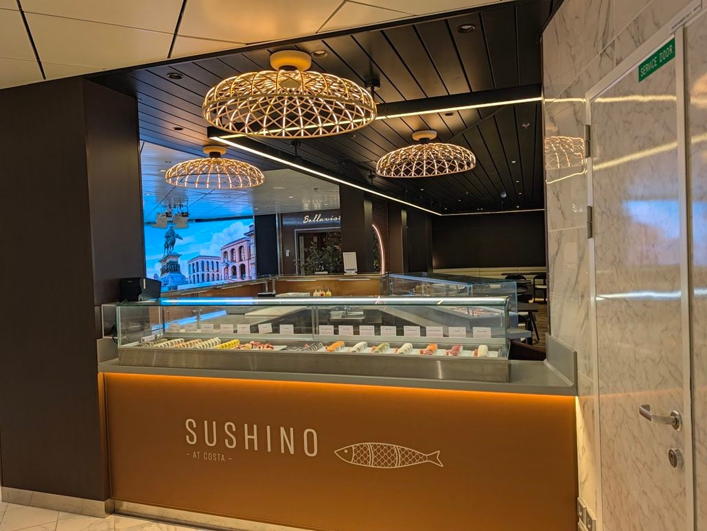 A sushi counter labeled “SUSHINO - AT COSTA” aboard the Costa Toscana, featuring a glass display case of sushi rolls illuminated by three round, lattice-patterned pendant lights.