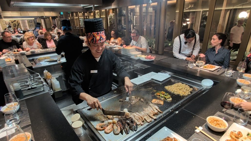 A chef in a black uniform and patterned hat prepares seafood and vegetables on a teppanyaki grill for customers seated at a Costa Toscana counter.