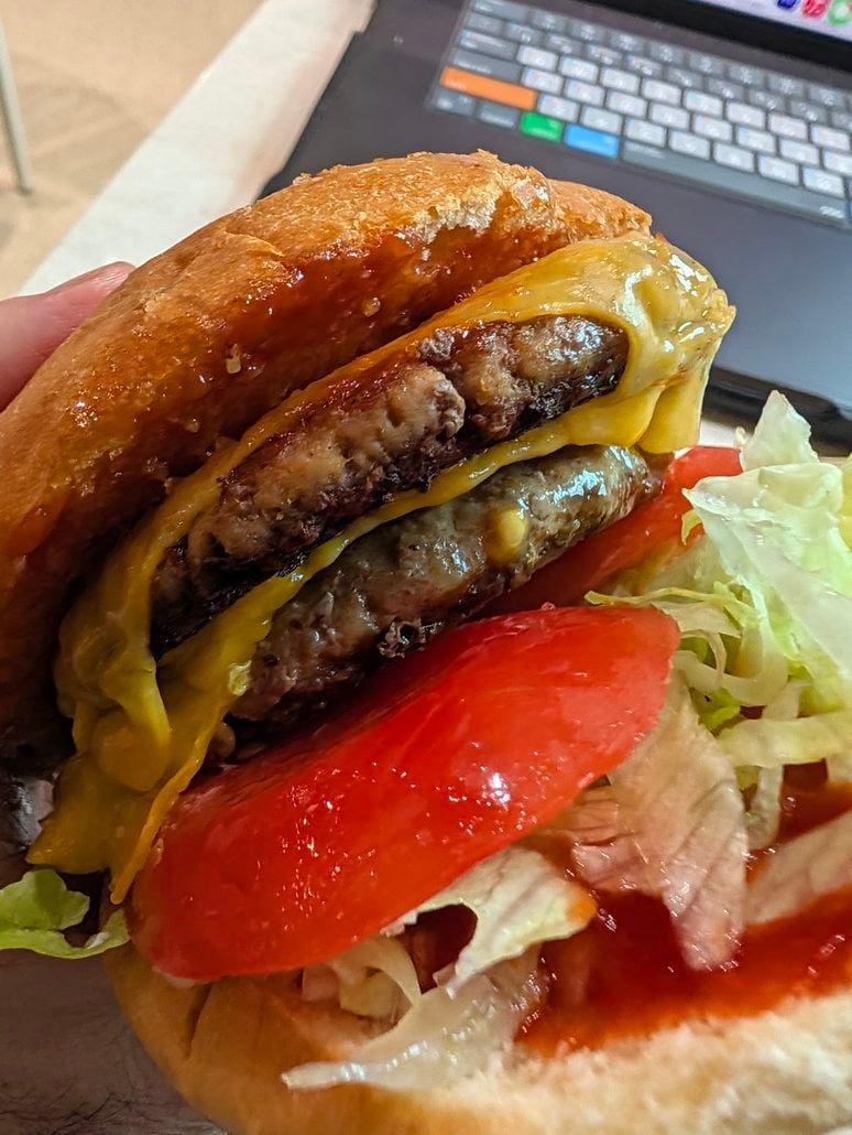 A double cheeseburger held in a hand, featuring lettuce, a tomato slice, and ketchup on a bun, with a laptop blurred in the background of a Costa Toscana cruise review.