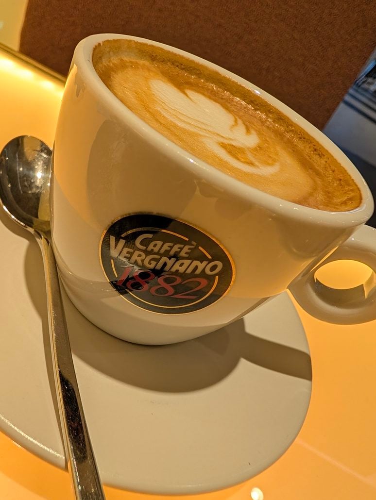 A cup of latte art coffee with a Caffe Vergnano 1882 logo on a saucer with a silver spoon, as enjoyed on the Costa Toscana.