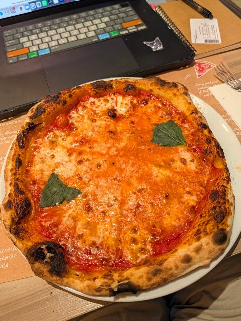 A cheese pizza with two basil leaves on a white plate next to a laptop during a Costa Toscana cruise review.