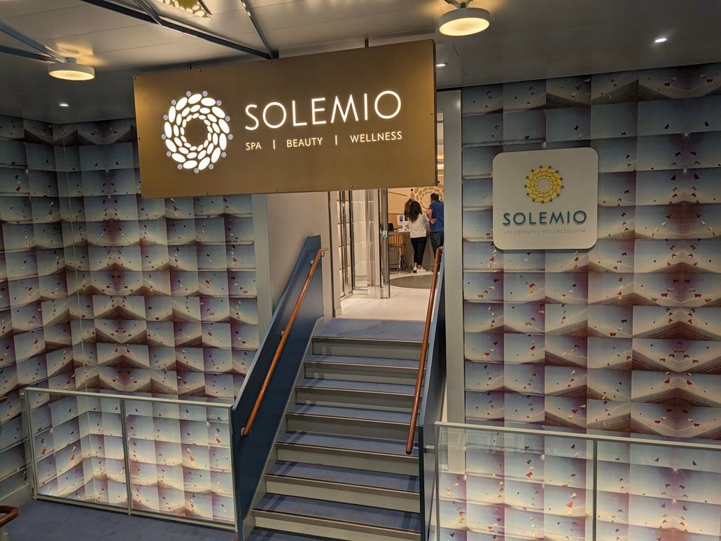 A short staircase leads up to the Solemio spa entrance on the Costa Toscana, featuring an illuminated sign and a wall patterned with a repeating abstract print.