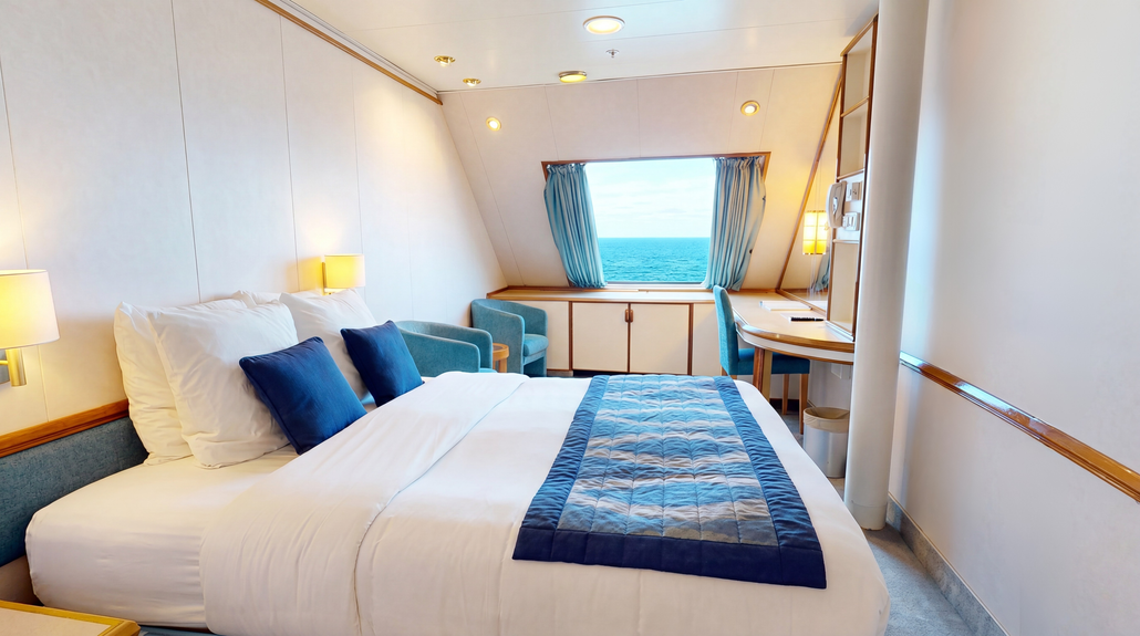 Cruise ship cabin with a double bed, blue accent pillows, a desk, and a window overlooking the ocean, ideal for full-time cruise living.