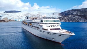 The Odyssey cruise ship sails on blue water in front of a glacier and rocky mountains, embodying the true cruise lifestyle.