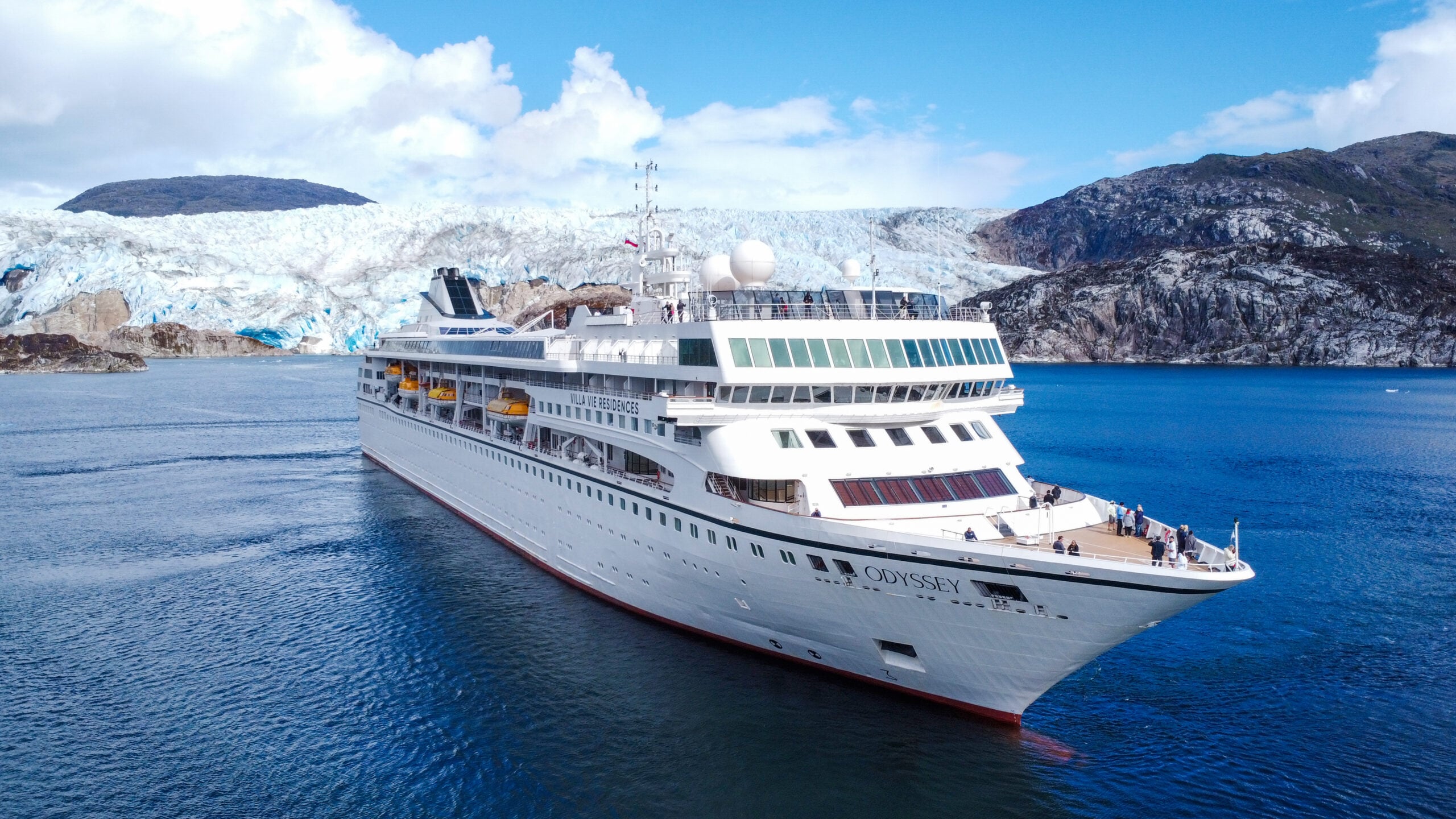 The Odyssey cruise ship sails on blue water in front of a glacier and rocky mountains, embodying the true cruise lifestyle.