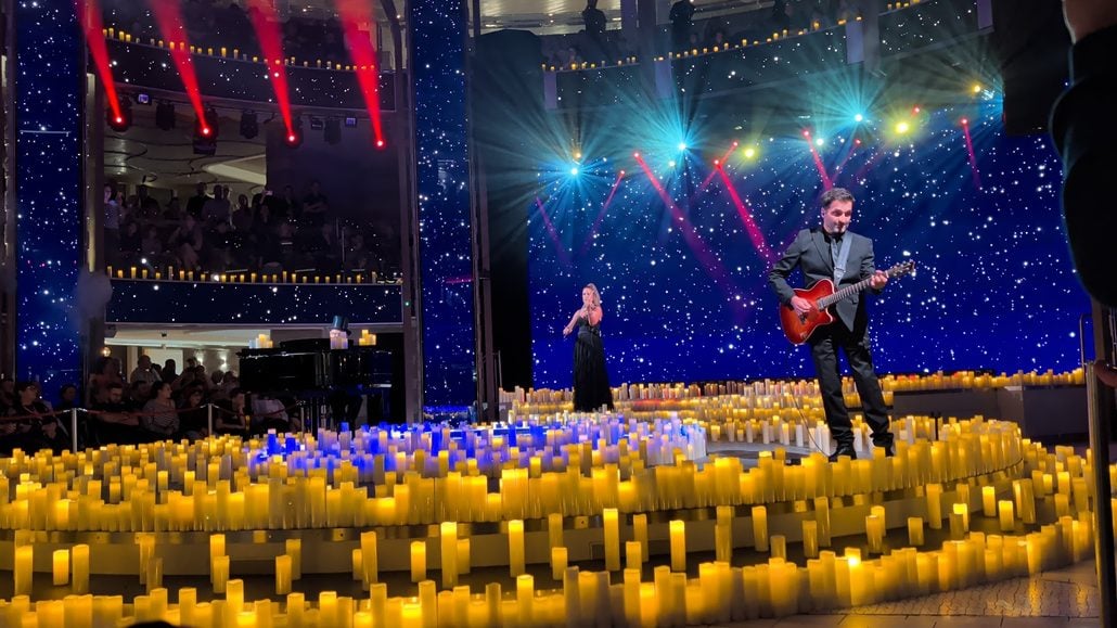 A woman playing the violin and a man playing an electric guitar perform on the Costa Toscana stage, surrounded by hundreds of lit candles in front of a blue star-patterned backdrop.