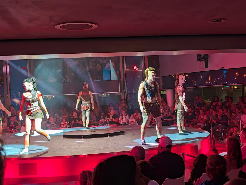 Four performers in tribal-style costumes and body paint stand on a circular stage aboard the Costa Toscana in front of a seated audience.
