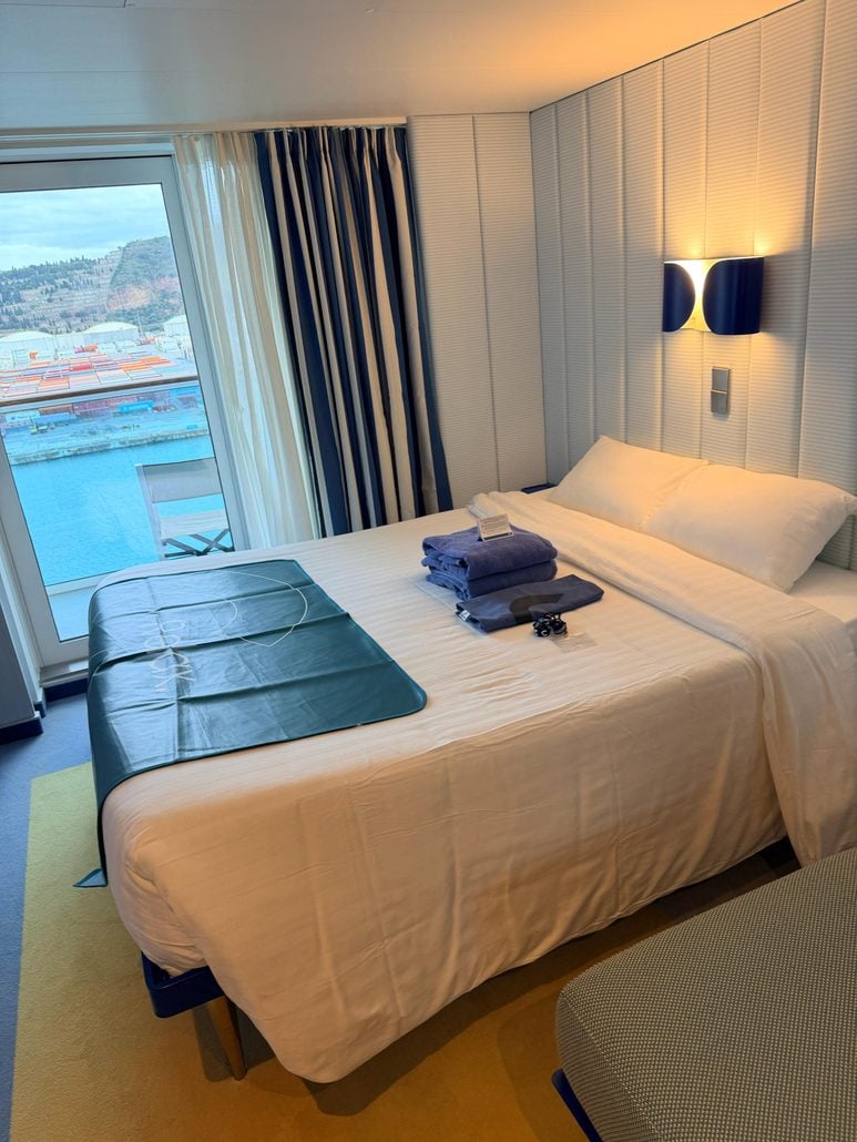 A Costa Toscana cabin with a made bed, folded blue towels, and a balcony view overlooking a shipping port.