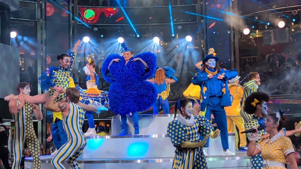 A large troupe of performers in colorful, patterned costumes dance on the multi-level stage of the Costa Toscana under bright spotlights.