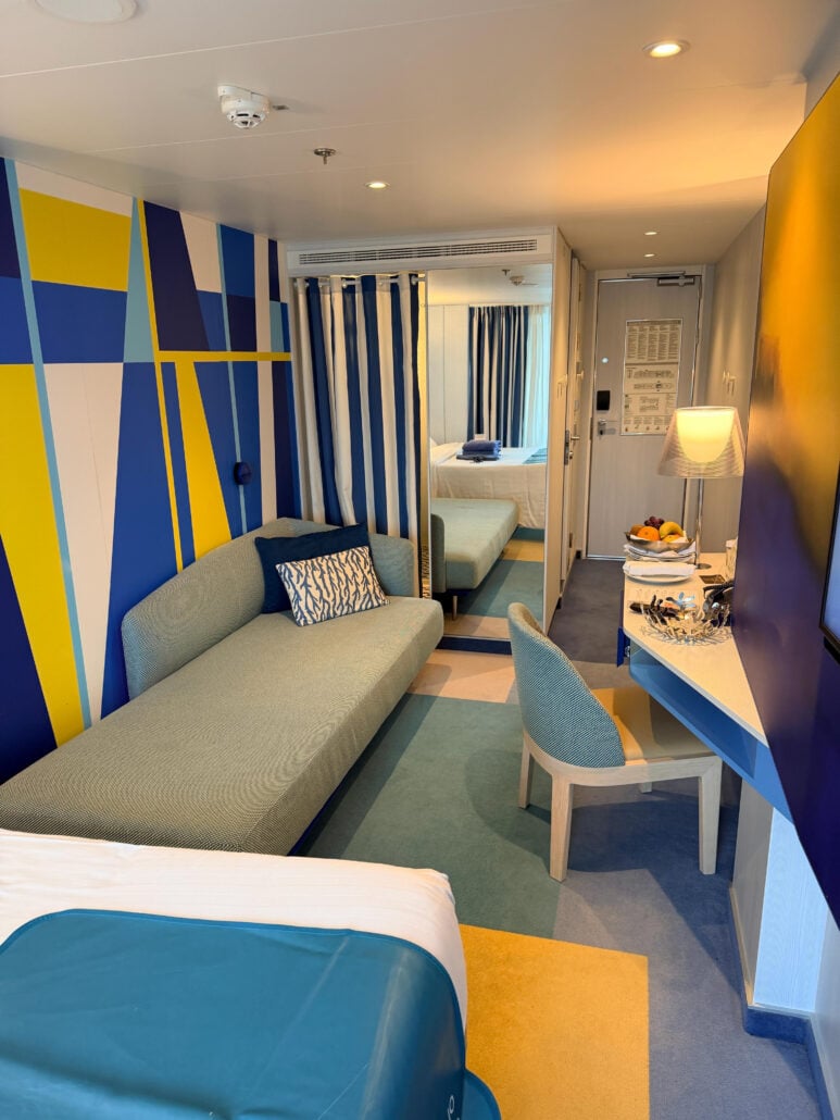 A Costa Toscana cruise ship cabin featuring a sofa, a vanity with a fruit bowl, and a mirror reflecting a bed with blue and white striped curtains.