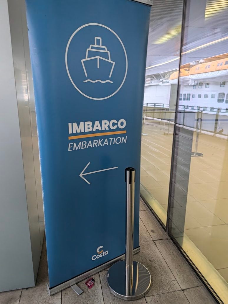 Blue Costa Toscana sign with a cruise ship icon and an arrow pointing left, labeled “IMBARCO” and “EMBARKATION.”
