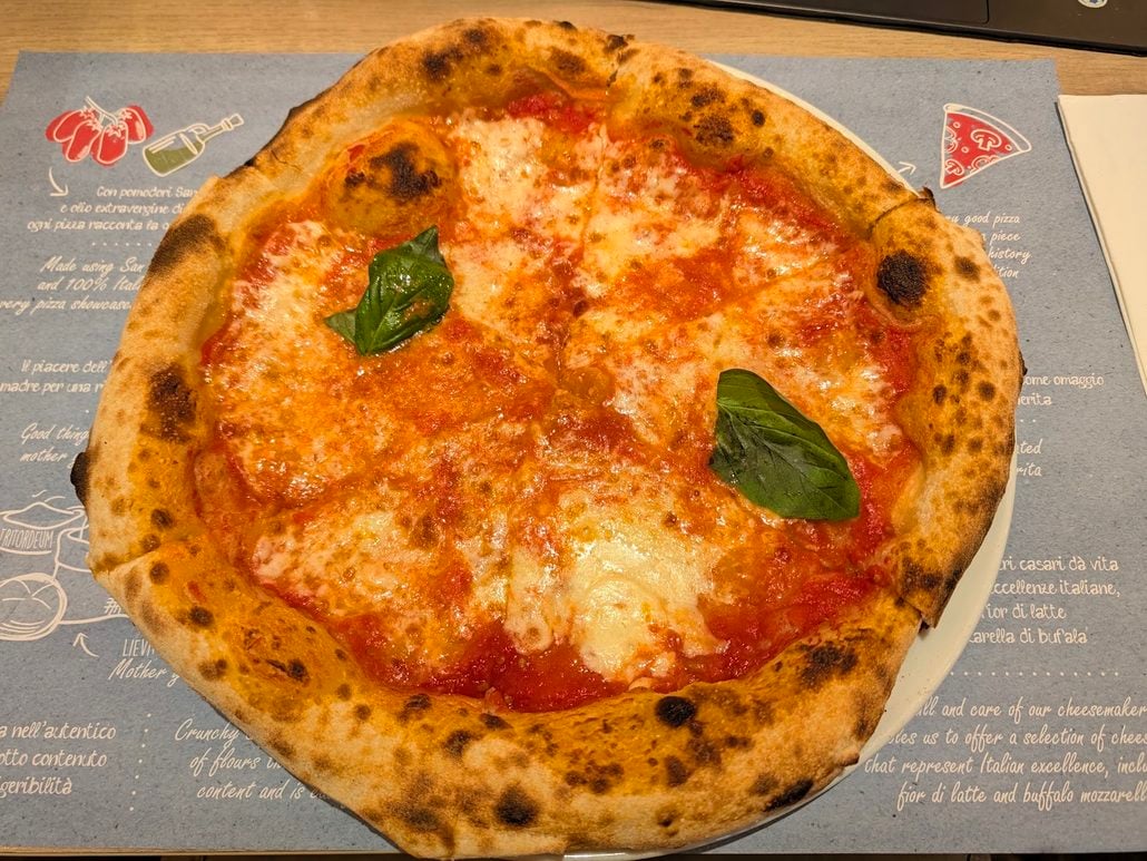 A Margherita pizza with tomato sauce, melted mozzarella, and two basil leaves on a Costa Toscana printed paper placemat.