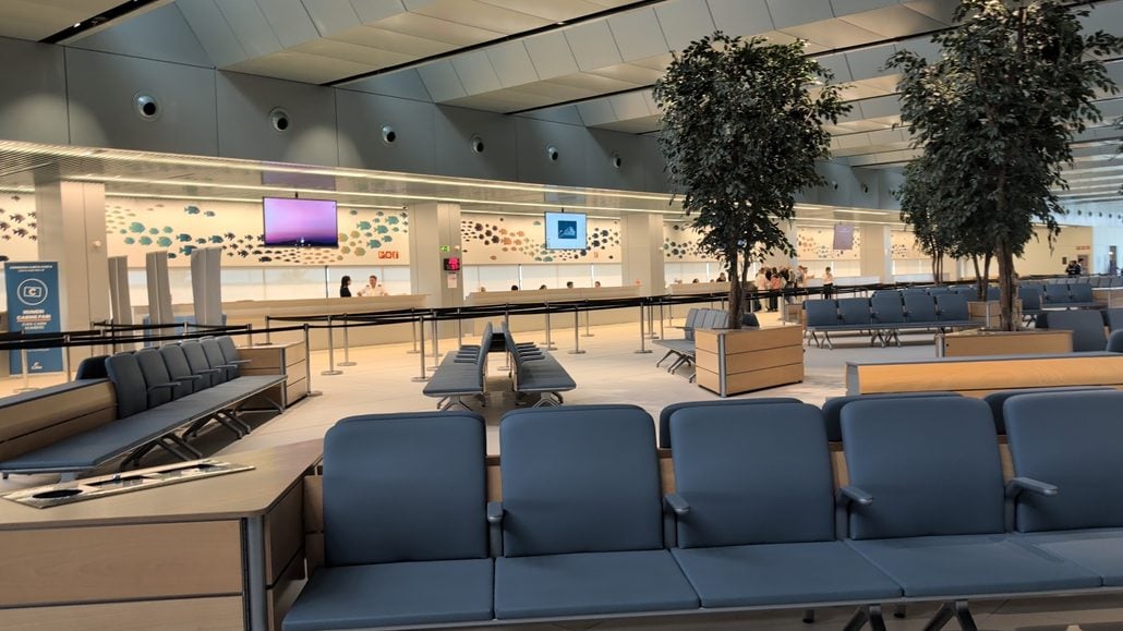 An airport terminal waiting area on the Costa Toscana with rows of blue chairs, two potted trees, and check-in counters in the background.