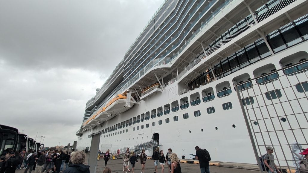 A large white Costa Toscana cruise ship docked at a port with a crowd of people walking along the pier.