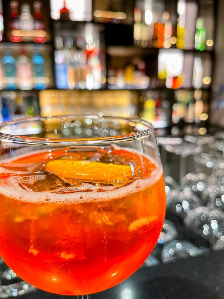 An orange spritz cocktail with ice and an orange slice in a wine glass, set against a blurred Costa Toscana bar background.