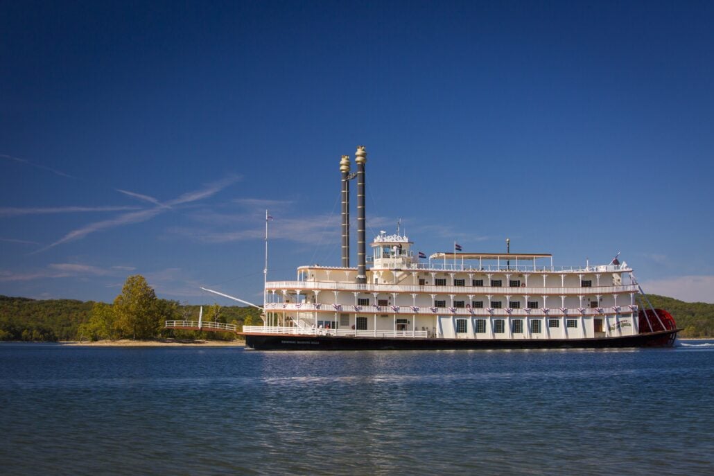 A large white paddlewheel riverboat cruises on a lake near a tree-lined shore under a clear blue sky, a classic sight for those planning a trip to Branson.
