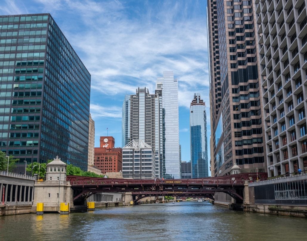 A red metal drawbridge spans the Chicago River, framed by skyscrapers in the Chicago Loop under a blue sky with light clouds.