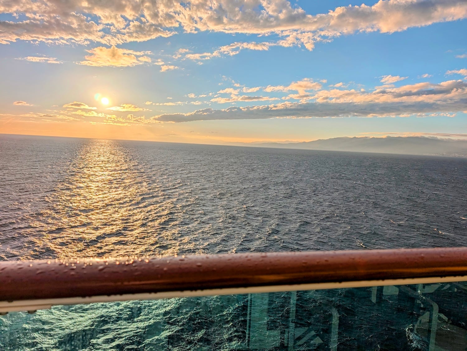 The sun sets over the ocean, viewed from a Costa Toscana balcony with a cruise port railing in the foreground.